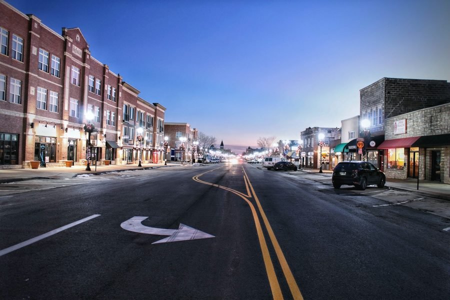 A small-town main street at dusk with brick storefronts lit and a car parked at the curb